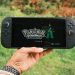 A hand holding a handheld gaming console displaying the Pokémon Legends game screen outdoors.